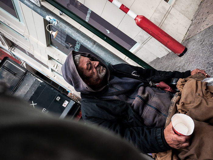 A shot from the hip tilted photograph of a homeless man in a wheelchair holding an empty cup in an urban street scene