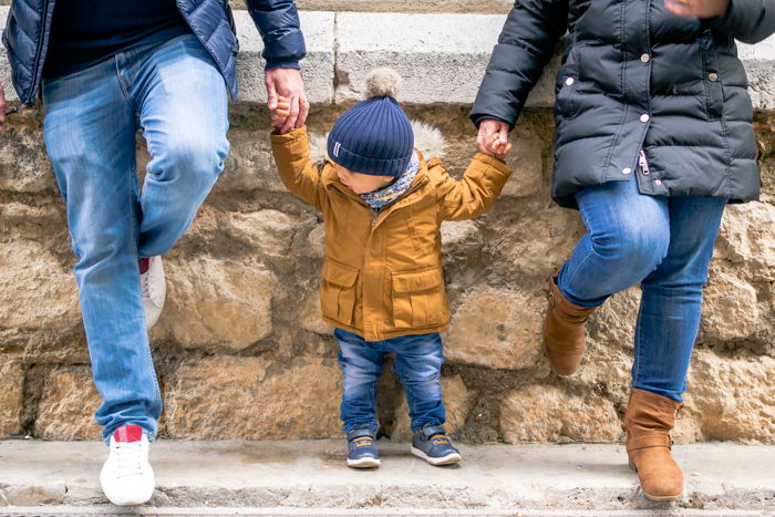 Family portrait photo of the legs of a couple holding their babys hands leaning against a wall