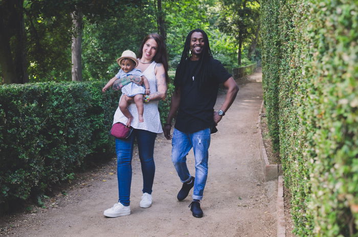 Family portrait photo of a couple and small baby walking outdoors by bushes and trees.