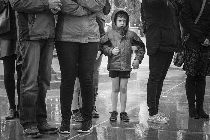 Black and white street photography of a little boy in the centre of group of adults in the rain. Street photography tips