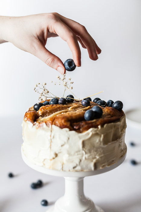 Scrumptious looking food photography shot of hand placing a blueberry on top of a cake against a white background, shot with a macro lens for still life photography