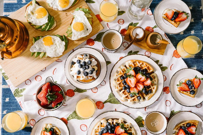 Scrumptious looking food photography overhead shot of a setup of many plates filled with fruit, pastries and eggs on a patterned tablecloth shot with a macro lens