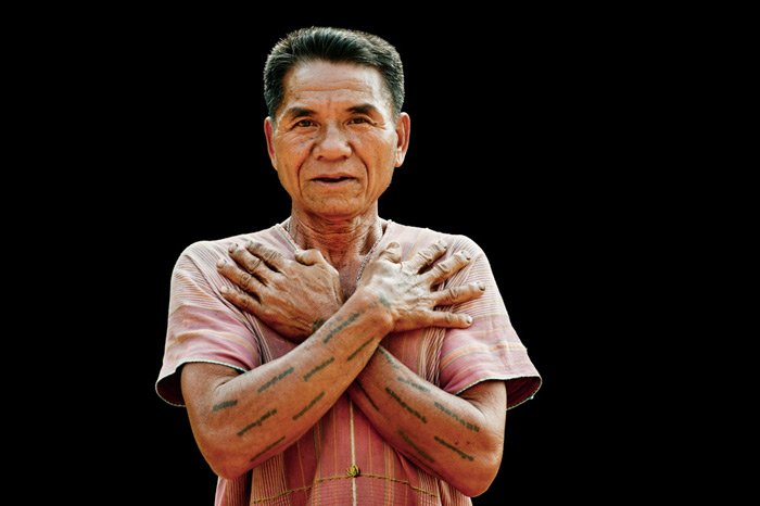 A Karen man shows off his traditional tattoos against a black ground in an outdoor portrait photography studio.