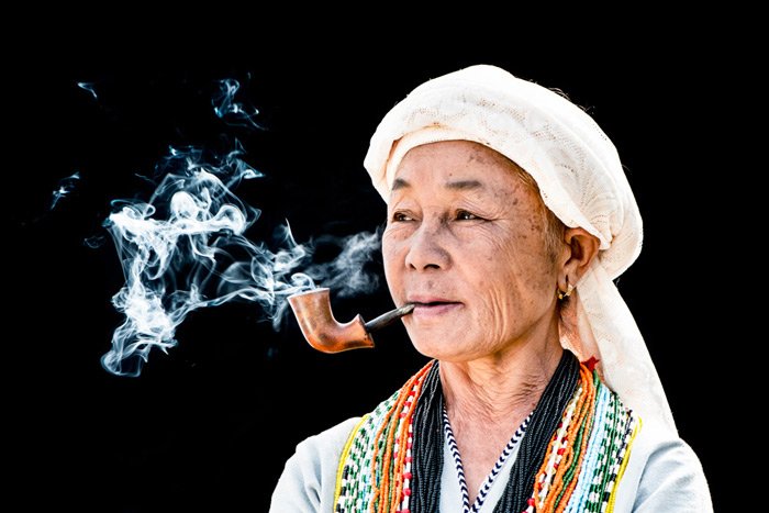 Photo of a Thai woman smoking a pipe against black backdrop in an outdoor portrait photography studio.