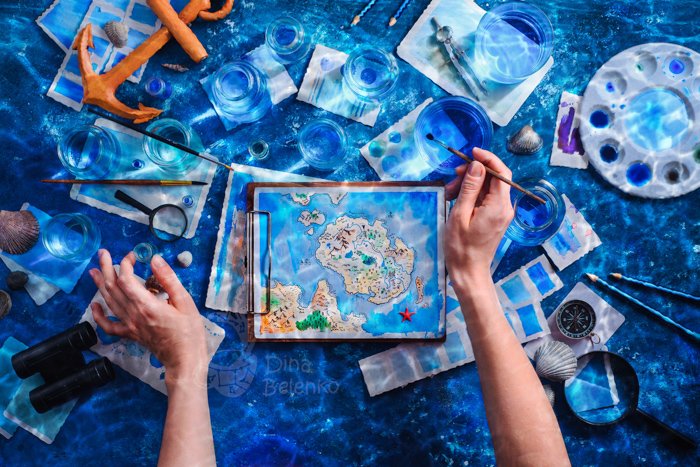 Overhead shot of an artist's hands painting a fantasy island map with watercolor paint and brushes in an underwater scene with an anchor and shells
