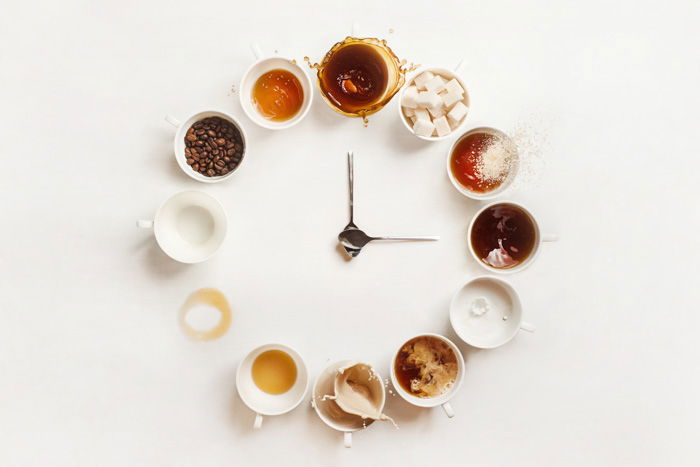 Overhead still life shot of a circle of coffee cups and two teaspoons in the center acting as clock hands