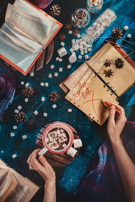 Overhead mystical shot of a person writing a 'to do' list in a notebook while holding a cup of coffee, sugarcubes, pine cones and other props are strewn around a glittery blue background