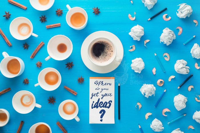 Overhead still life of coffee cups, cinnamon sticks, balls of paper and pencils with a notepad with the words 'where do you get your ideas' on sky blue background