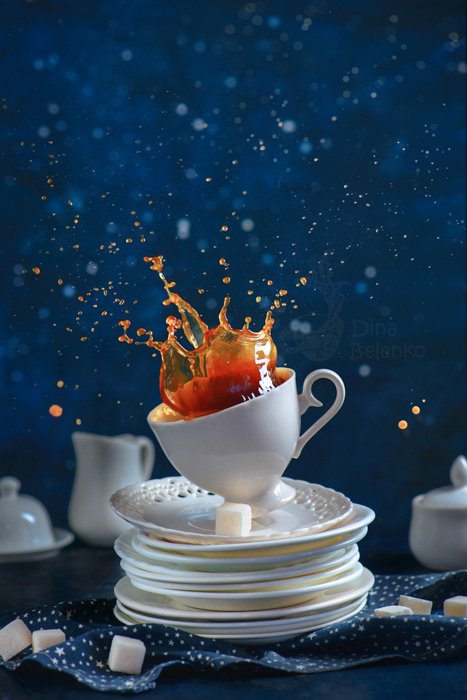 A playful food photography shot of a coffee-cup with splashes balancing on a pile of white saucers against a blue background