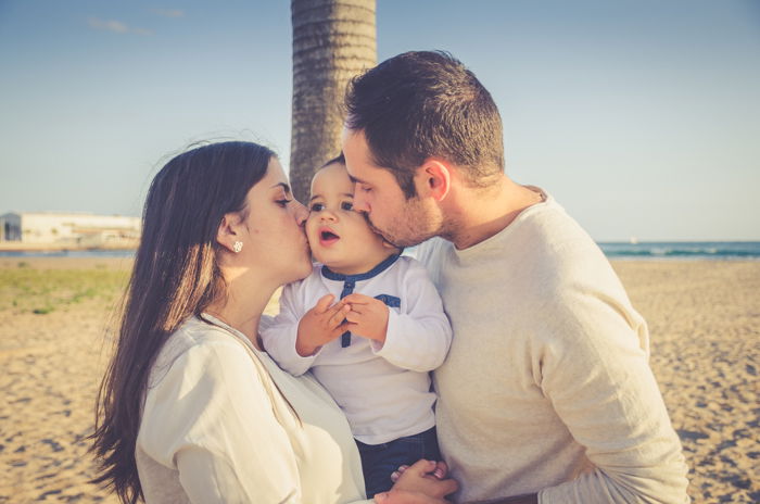 Sweet portrait of a man and woman holding a small baby and kissing his cheeks on a beach - family portraits composition