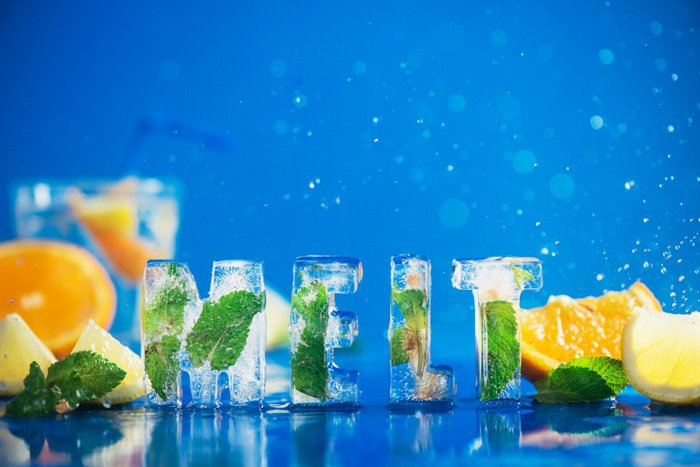 Food typography: Ice cube lettering with frozen mint leaves, lemon slices and oranges on a blue background with water splashes. Text says Melt. Water drop bokeh. Copy space.