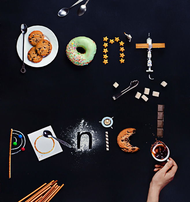 An overhead shot featuring coffee cups, cutlery, toys,saucers, biscuits and other food art props with food typography message 'don't panic'