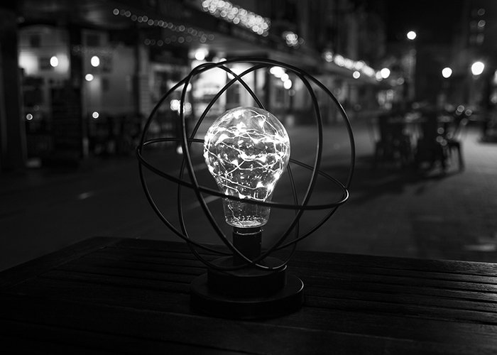 A black and white street shot of a lightbulb sculpture on a wooden table at night