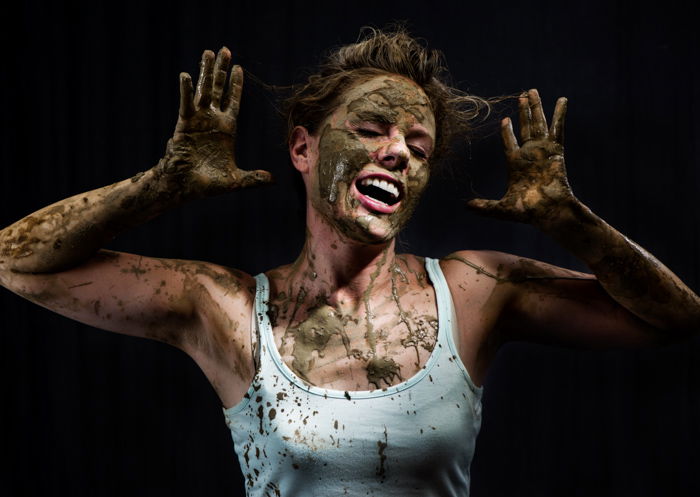 Portrait of a woman covered in ceramic clay with hands by her head again a dark background