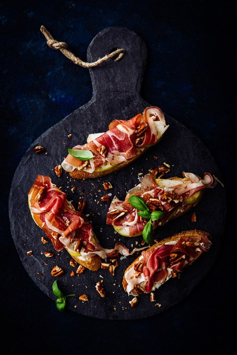 Overhead shot of prosciutto crostini with pecans in a dark food photography style