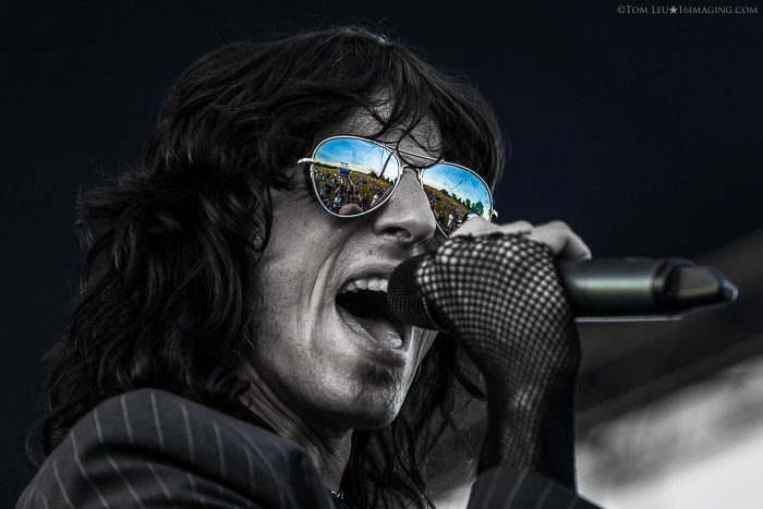 A black and white concert photography live shot of a singer onstage, the crowd are reflected in clour in his sunglasses