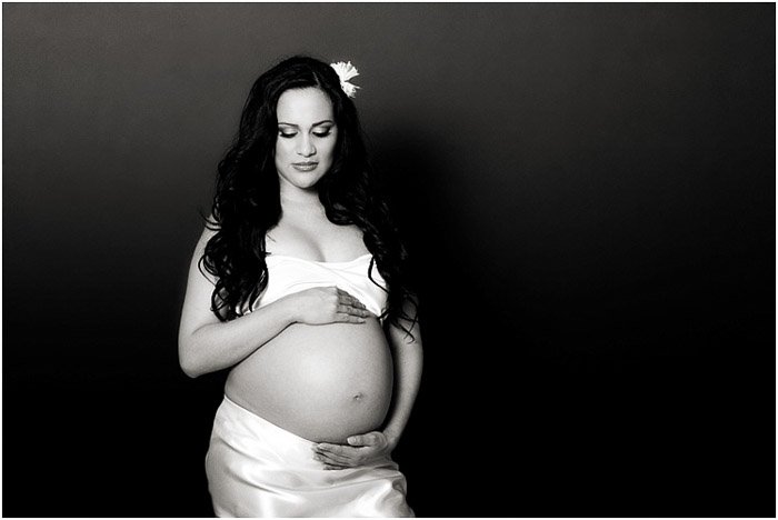 A black-and-white photo of a pregnant woman with her belly exposed and her hands around her stomach as a maternity pose