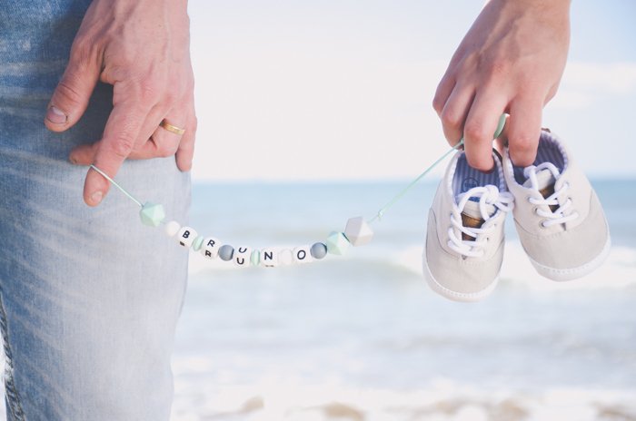 Close-up of a person holding a baby's shoes and name necklace on a beach photoshoot