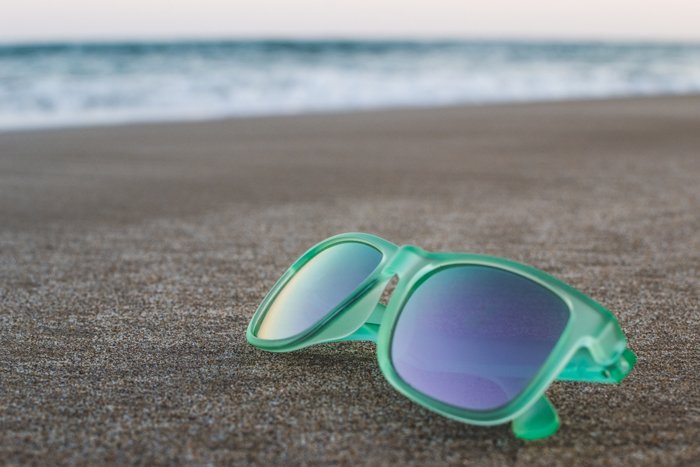 A pair of green framed sunglasses resting on the sand, the seas waves blurred in the background