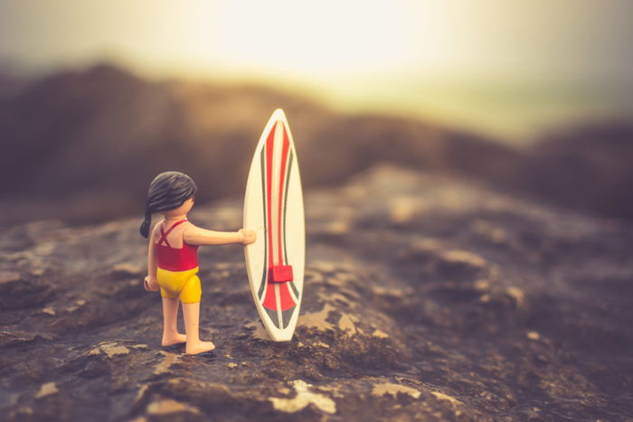 A Playmobil character with a surfboard posed on a rocky beach