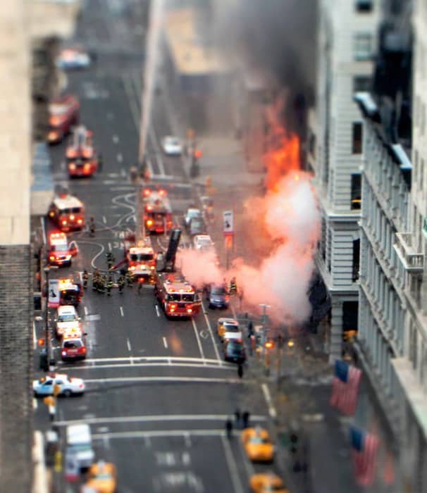  Overhead shot of a fire in an urban setting. Fire engines and fireman and many cars line the streets. The use of tilt shift photography makes the scene look like its made from toys.