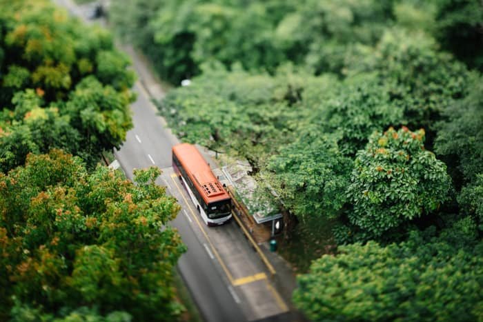 A tilt shift photo of a bus parked on a road in a beautiful countryside area surrounded by trees