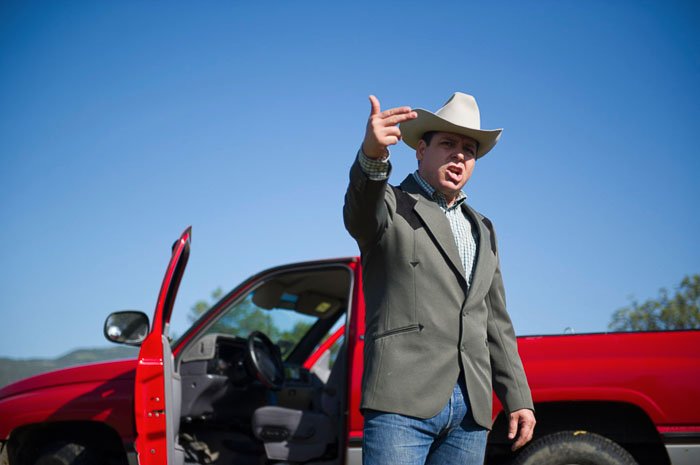 A man in a cowboy hat standing in front of a red car and making a hand gesture - tipping for photos of people