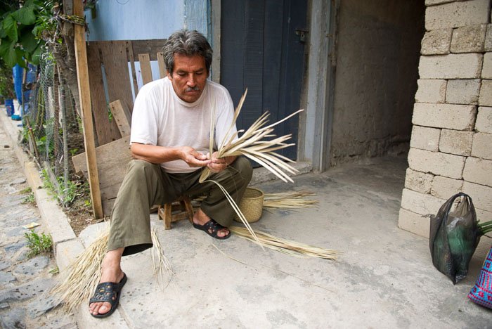 A travel photo of a man sitting outside a stone building, holding straw. 