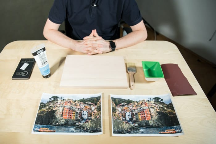 A man sits at a wooden table, before him is two identical photos of a brightly colored coastal town,glue, paintbrush, sandpaper and other materials to transfer laser print to wood