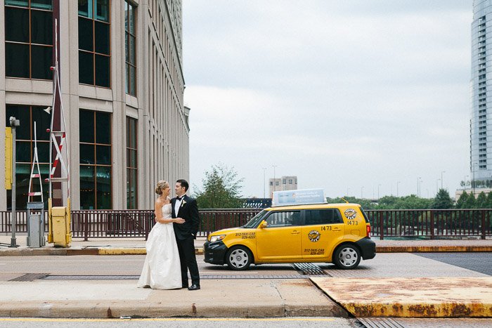 a newly wedding couple pose on the street in front of a yellow taxi 