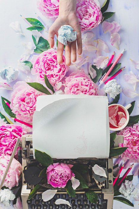 A bright and airy flat lay still life with a typewriter, pink roses and a hand holding a crumpled ball of paper