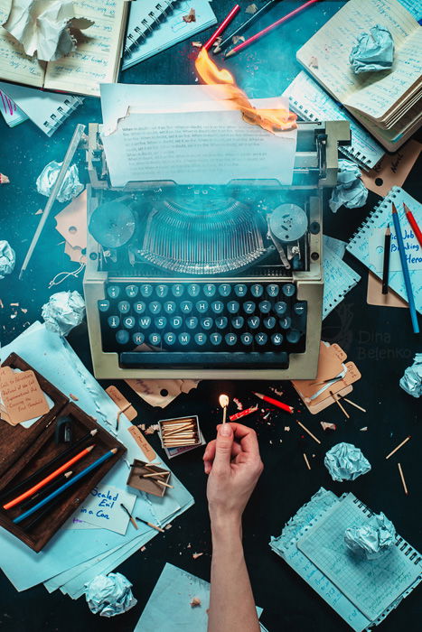 Atmospheric flat lay still life with a typewriter with burning paper surrounded by writing materials and a hand holding a lighted match 