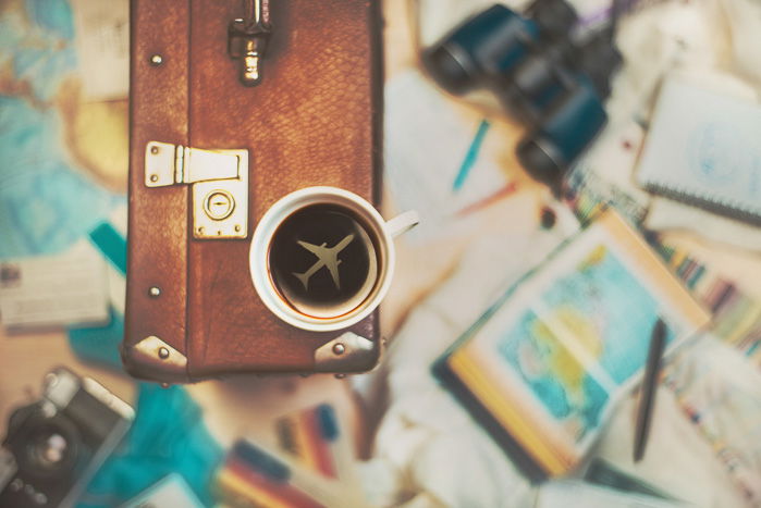 A travel themed still life flat lay with a cup of coffee perched on a suitcase with the shape of an airplane inside