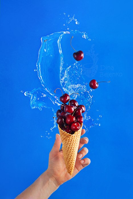 A hand holding an ice-cream cone filled with cherries against a light blue backdrop for flat lay photo