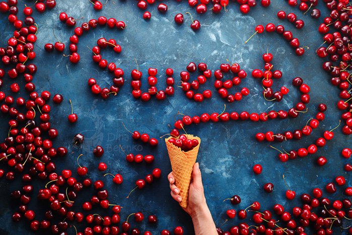 Flat lay photo idea: A fun food photo of cherries spelling the word 'sweet' on blue surface with a hand holding an ice-cream cone underneath