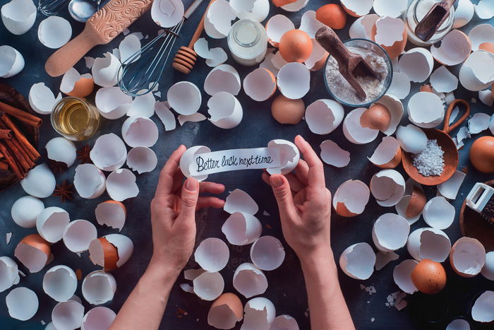 A food photography themed flat lay still life with egg shells, cooking utensils and hands holding a note saying 'better luck next time'