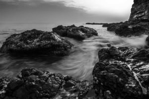 A black and white fine art photography shot of a rocky beach, with a soft misty effect of the water around the rocks and cliffs