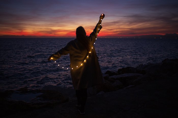 A person standing and facing the ocean, holding fairy lights behind them on a beach a sunset