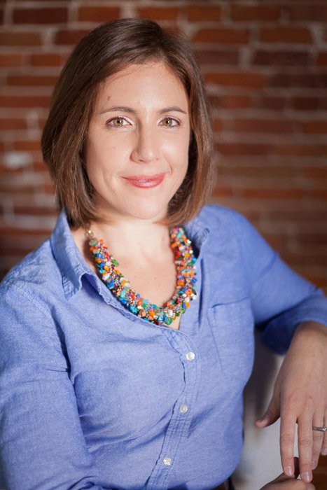 A corporate headshot of a woman in a purple shirt against a brick wall background
