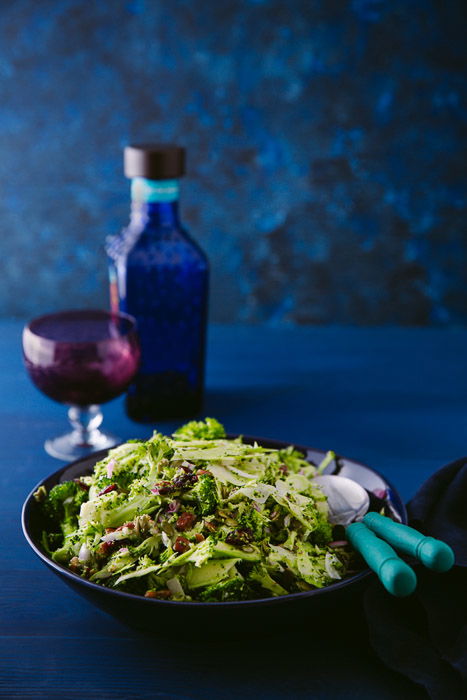 A bowl of shaved brocolli salad, balsamic veingear and a small glass on a dark blue background