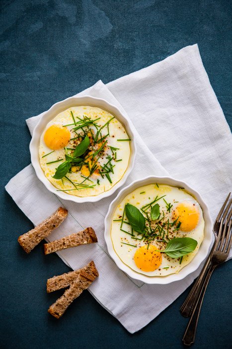 Overhead food styling shot of baked eggs in two white bowls against a dark blue background