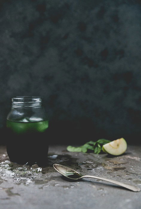 A still life featuring a green drink in a jar beside a teaspoon and ingredients against a dark background