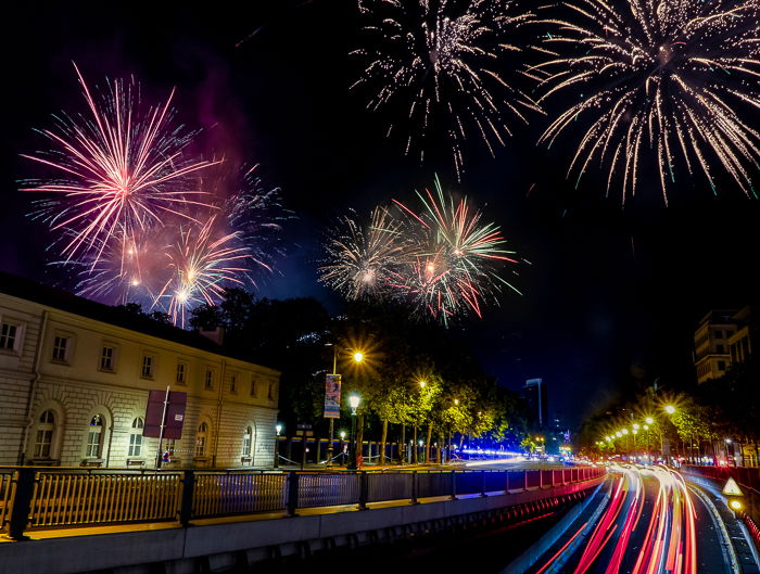 an urban street and building lit by colorful fireworks and light trails from vehicles on the street against a dark night sky