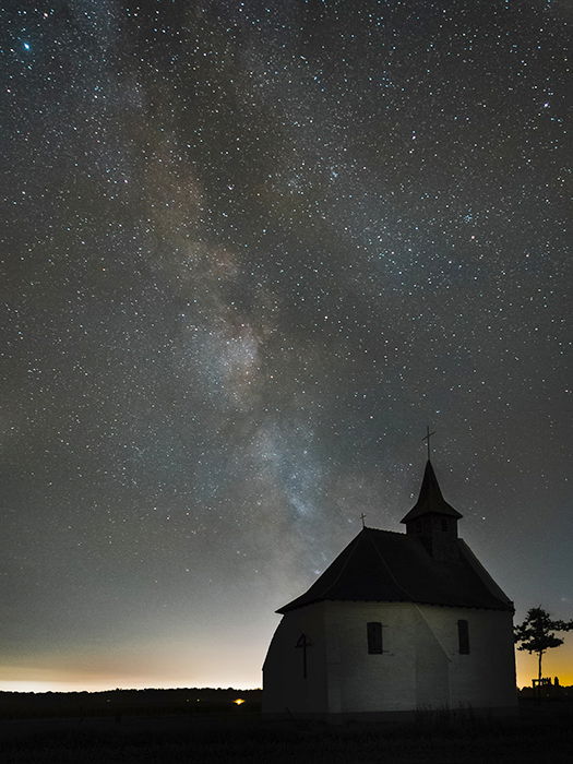 a chapel on a hill lit warmly from behind, against the night sky with the milky way visible