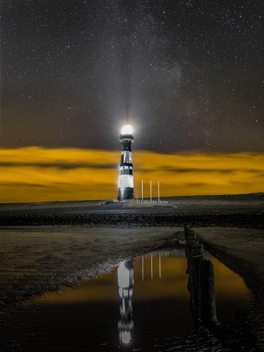 a lighthouse in the netherlands, yellow lit clouds in the background, reflected in a pool of water on the beach