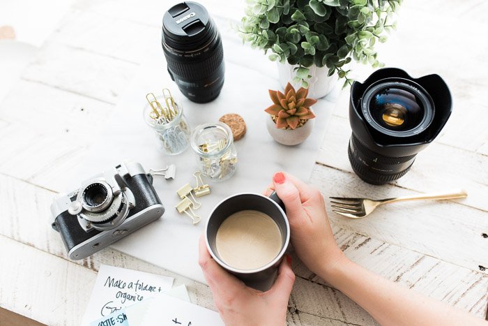 Overhead shot of a person holding a coffee cup at a table with plants, camera, lenses and other photography equipment