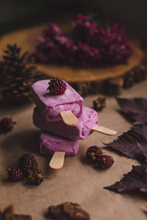 A dark and moody food photography shot of three pink ice cream lollys in a pile on a wooden surface with berries and leaves