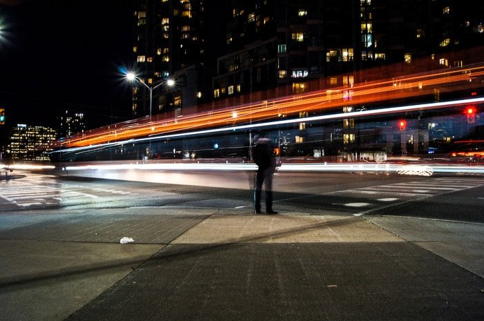 A man standing on a street at night, a stream of light trails in front of him