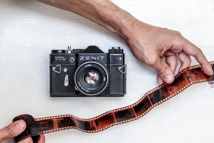 An overhead shot of a Zenit film camera on white background with a person holding a roll of film negative beside it - using film for street photography 