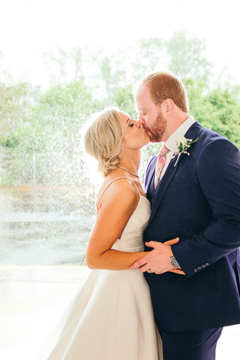 A wedding portrait of the bride and groom kissing outdoors in front of a fountain